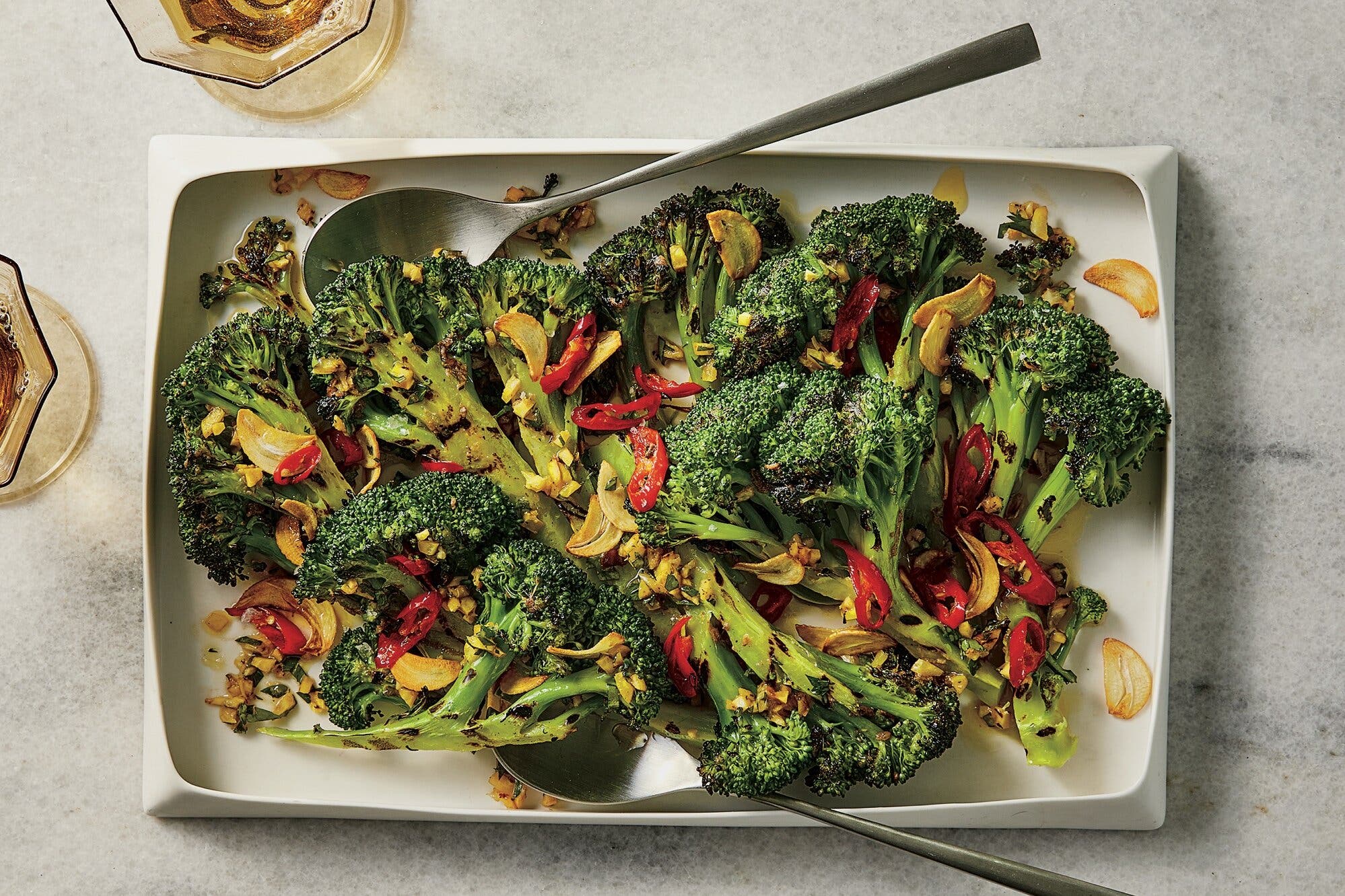 Broccoli topped with sliced garlic and pepper sits on a rectangular platter with a large metal spoon.