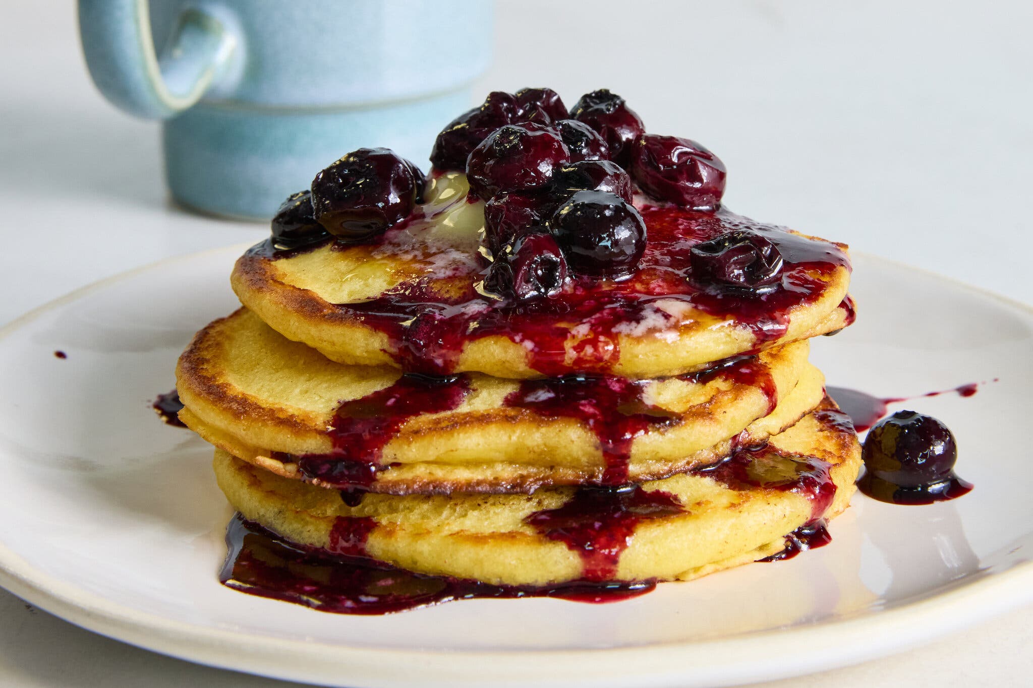 A stack of lemon ricotta pancakes topped with berry compote sits on a plate. A blue mug is visible in the backgroun.