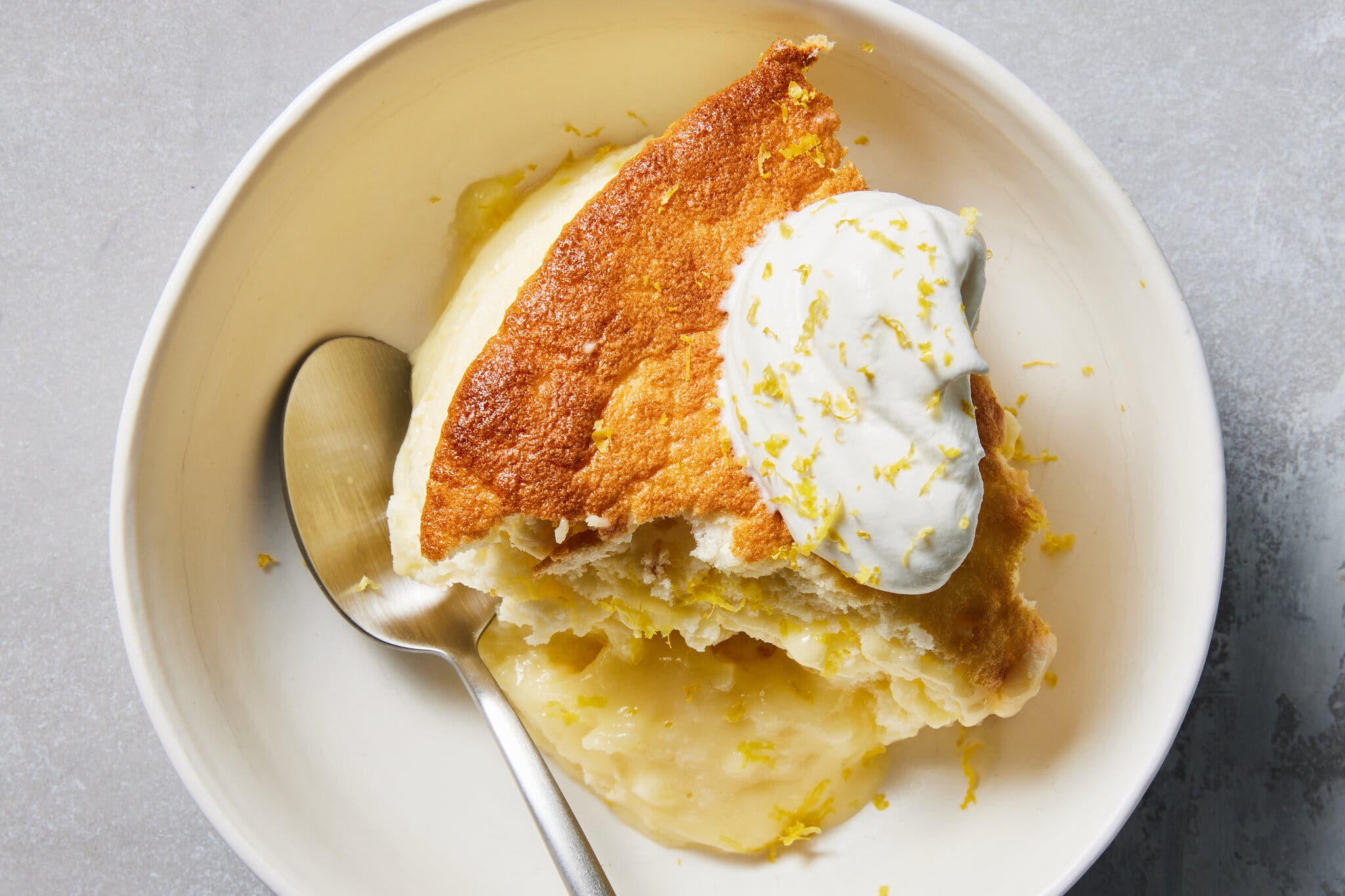 A slice of lemon pudding cake sits in a white bowl with a spoon.