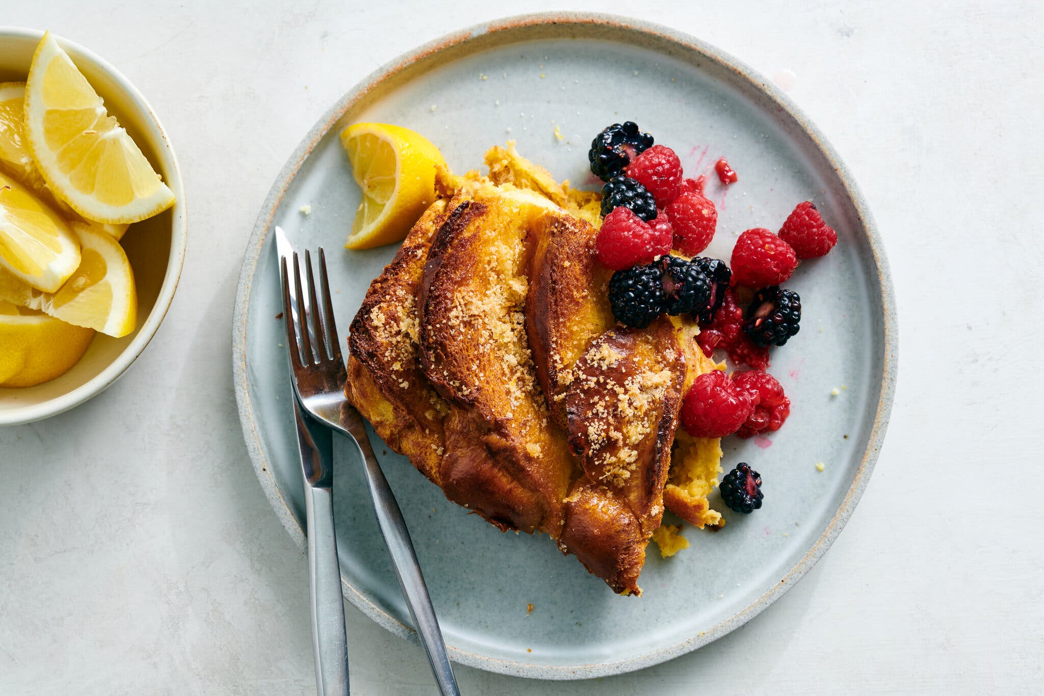 A square of French toast sits on a plate next to a lemon wedge and berries.