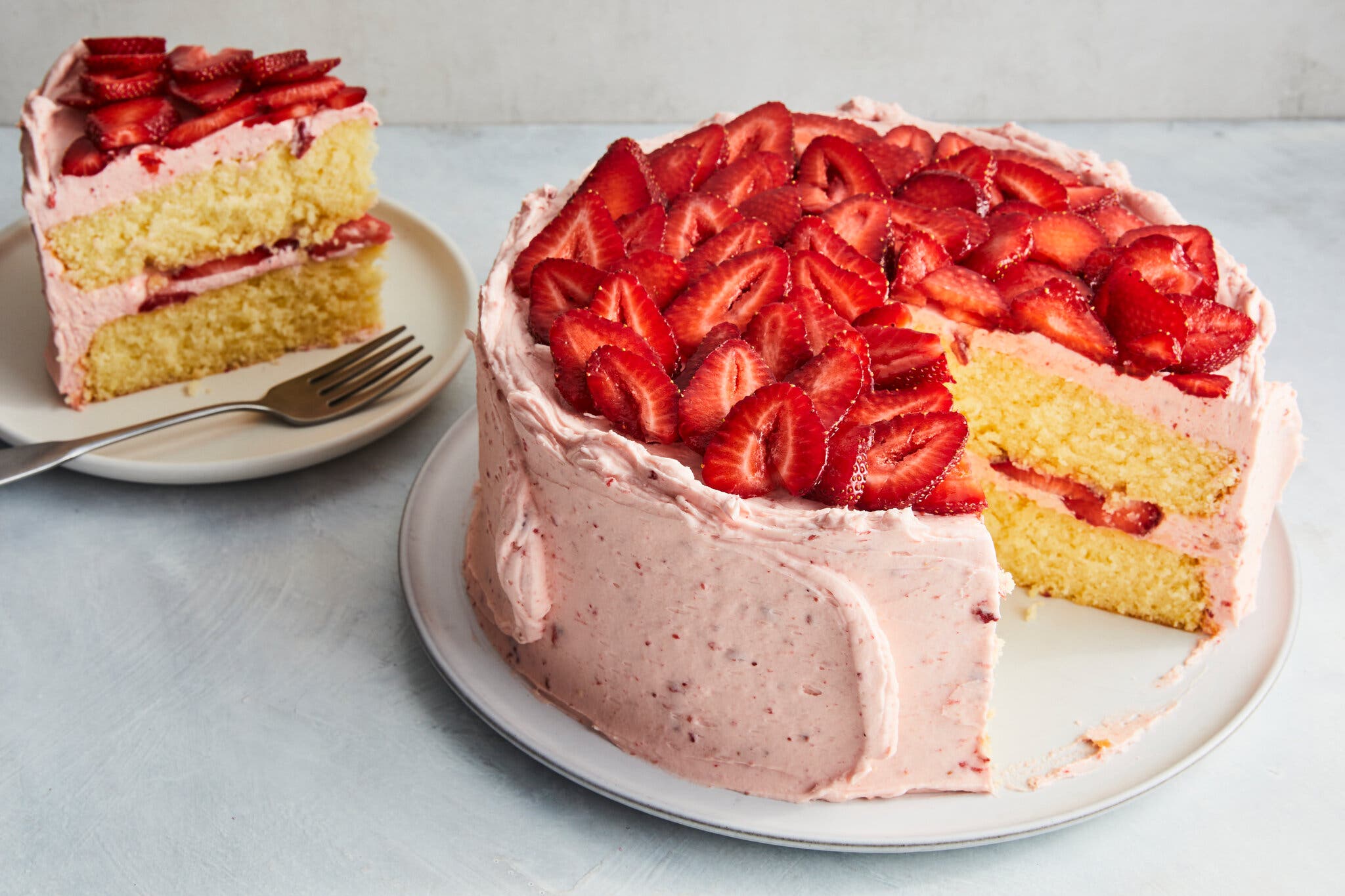 A slice of strawberry lemonade cake with pink frosting and topped with sliced strawberries sits next to the whole cake.