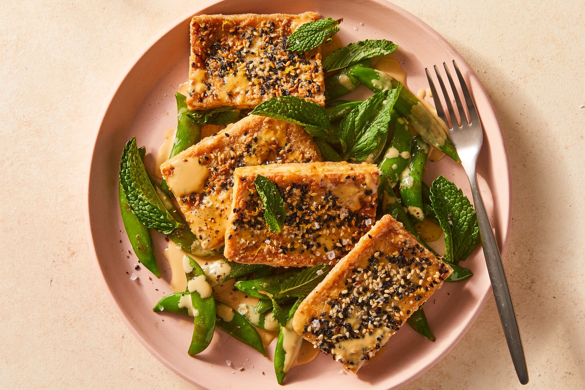 Crispy tofu topped with black and white sesame seeds sits on a pink plate surrounded by mint leaves and snap peas.
