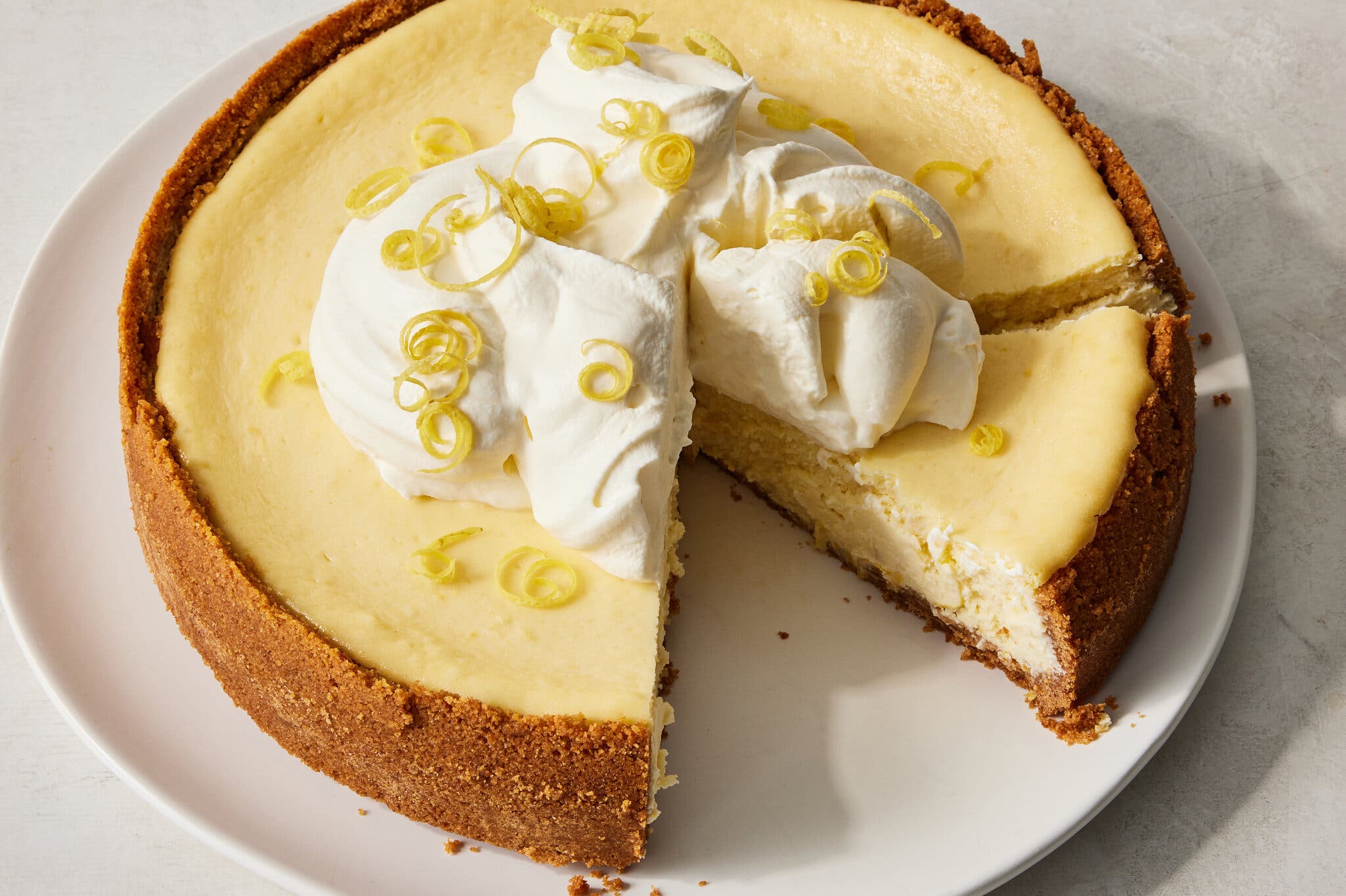 A lemon yogurt cheesecake with a slice taken out and another cut slice sits on a plate. It’s topped with whipped cream.