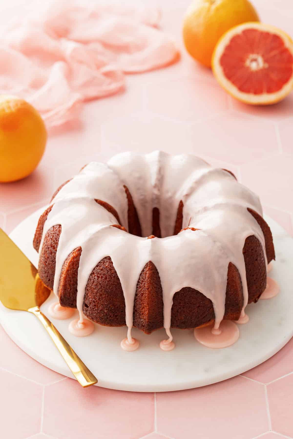 Pink glazed grapefruit bundt cake on a marble cake plate with a pink background and a cut pink grapefruit in the background.