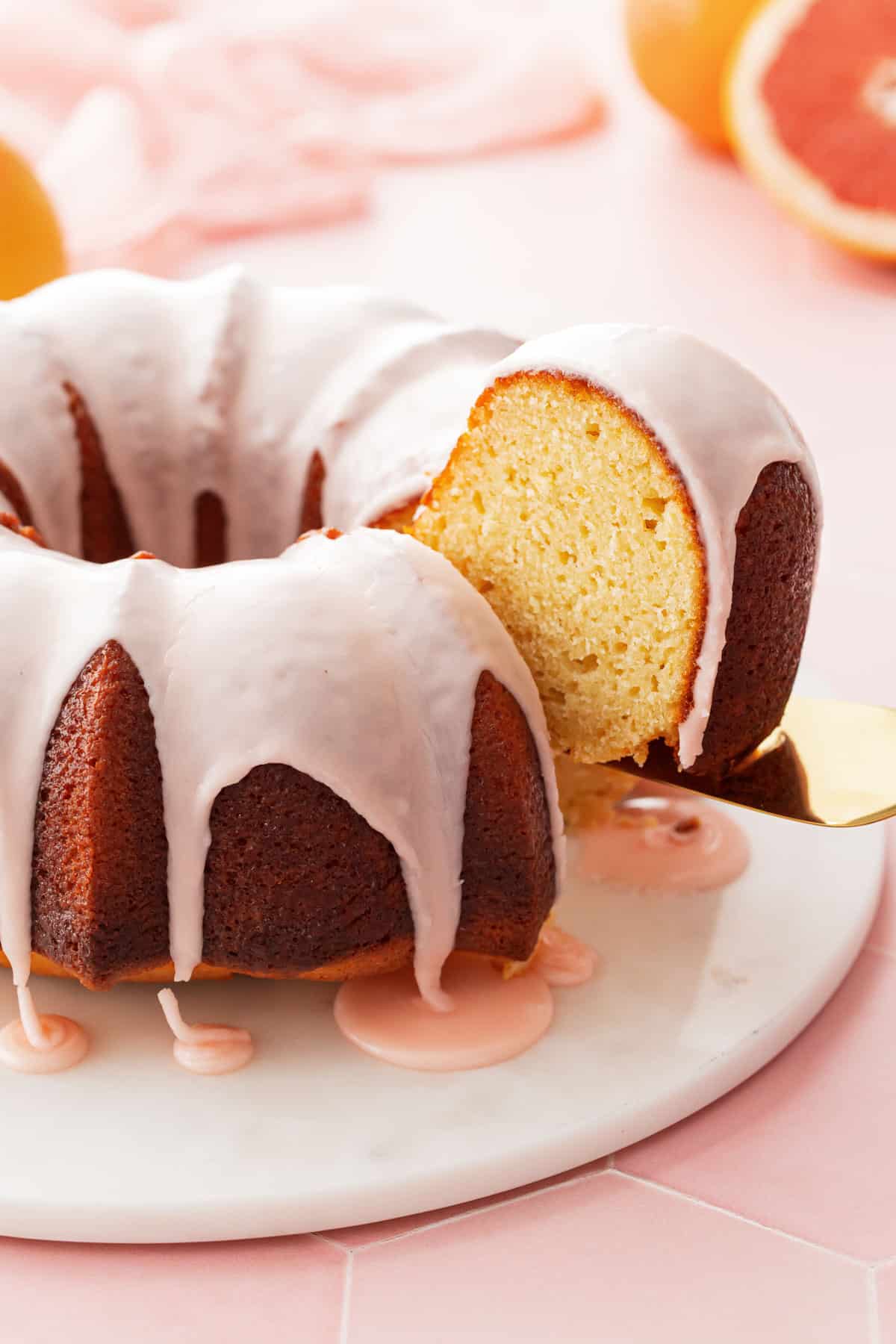 Cake server lifting a slice of Grapefruit Olive Oil Bundt Cake, sitting on a marble cake plate with drizzled glaze pools.