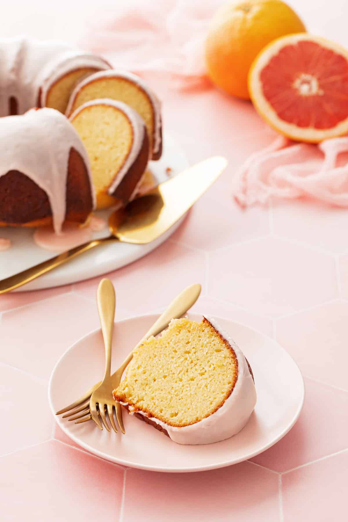 Pink plate with a slice of Grapefruit Olive Oil Bundt Cake and two gold forks, the rest of the cake and a cut grapefruit in the background.