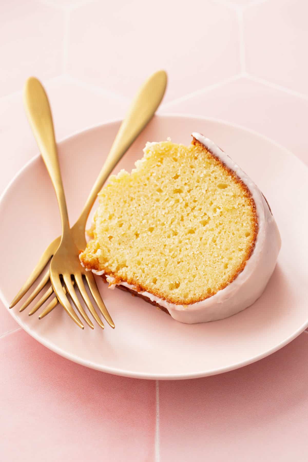 Closeup, slice of glazed grapefruit olive oil bundt cake on a pink dessert plate with two gold forks and a pink background.