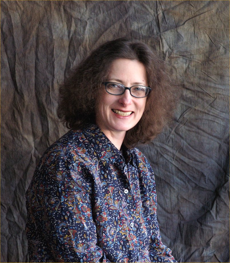 A woman with shoulder-length curly hair and glasses smiles at the camera. She is wearing a patterned shirt and is posed in front of a textured, brownish-gray backdrop.