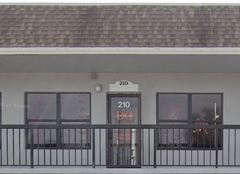 A small commercial building with a shingled roof and gray exterior. The door, labeled "210," is flanked by two large windows. A black railing runs along the front of the building.