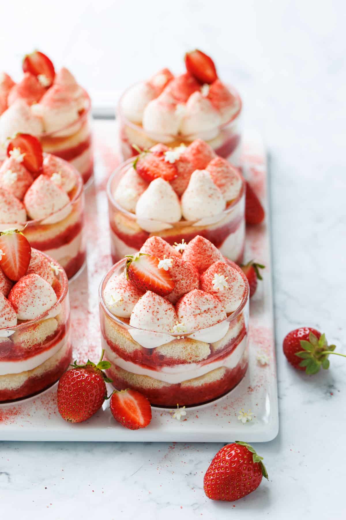 Rows of Strawberry Elderflower Tiramisu in clear ramekins on a white serving platter, decorated with freeze dried strawberry powder and elderflower blossoms.