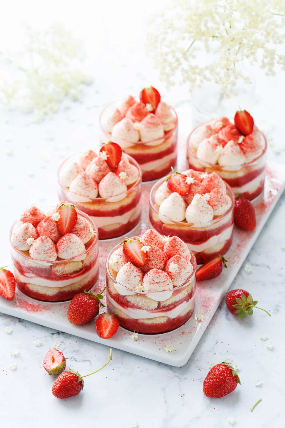 Strawberry Elderflower Tiramisu in individual clear ramekins, with fresh strawberries and a spray of elderflowers in the background.