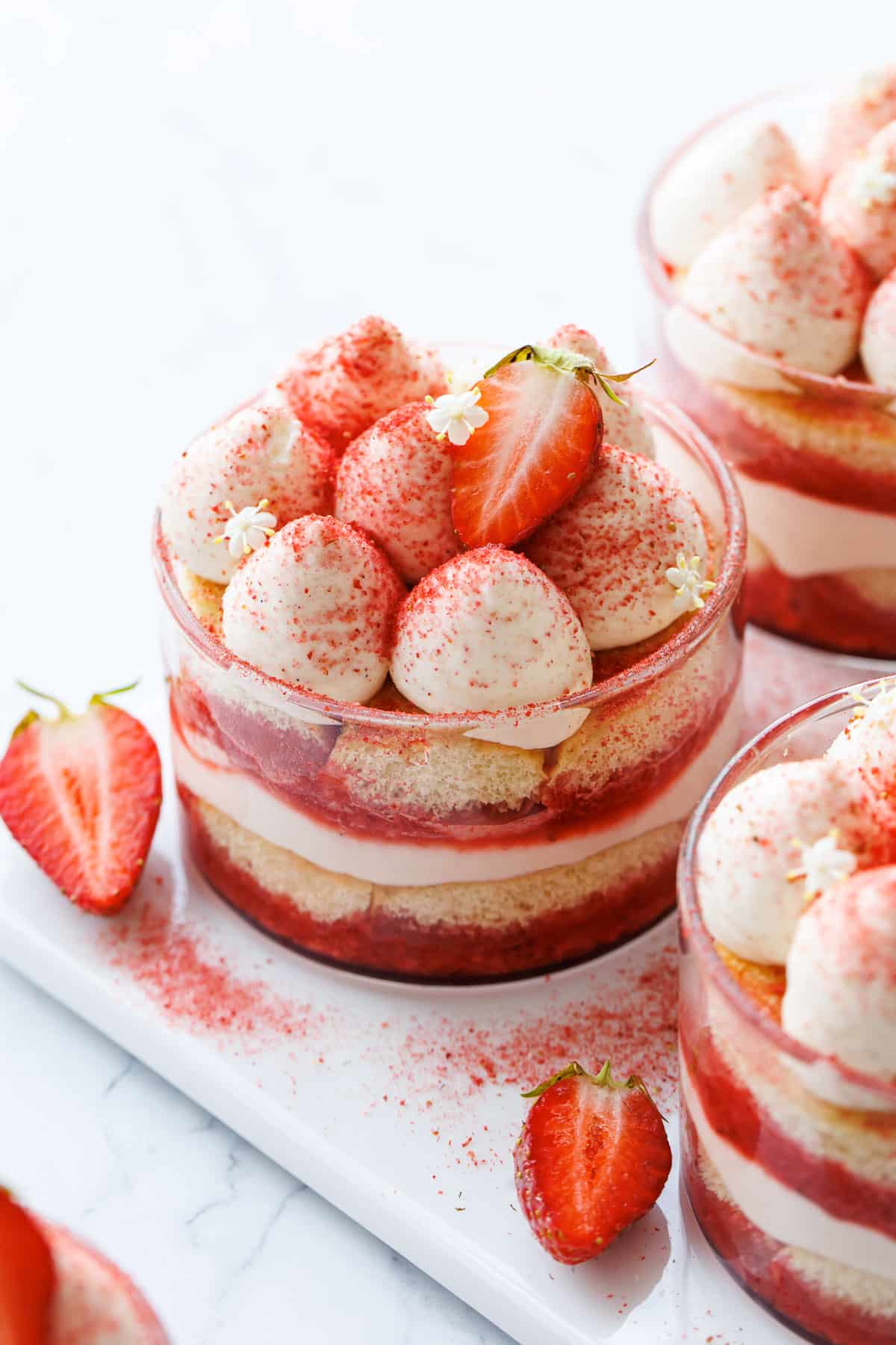 Strawberry Elderflower Tiramisu in individual clear ramekins, topped with freeze dried strawberry powder, a fresh strawberry cut in half, and a few elderflower blossoms.