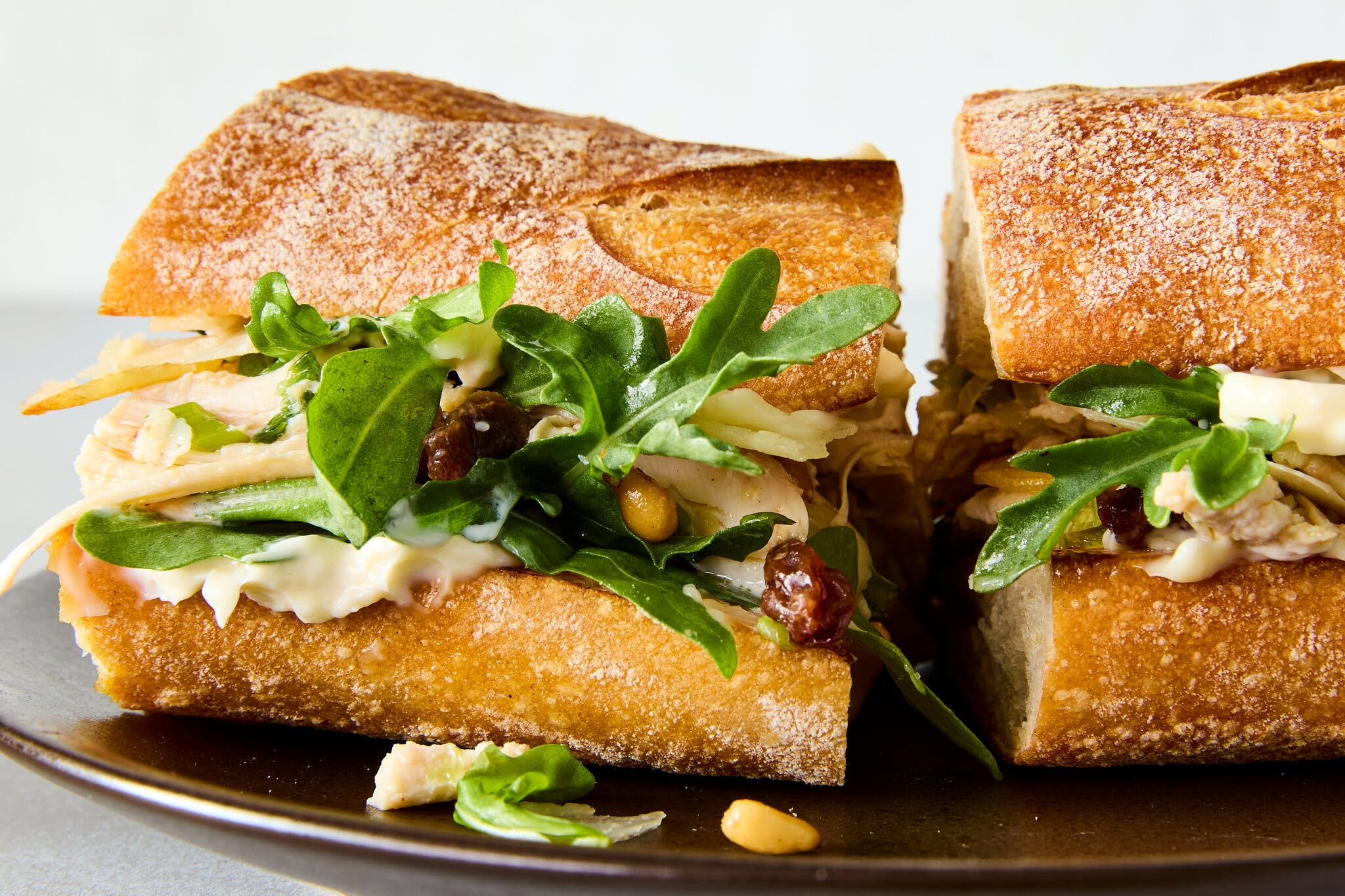 A chicken sandwich on a baguette, filled with arugula and currants, is photographed from the side.