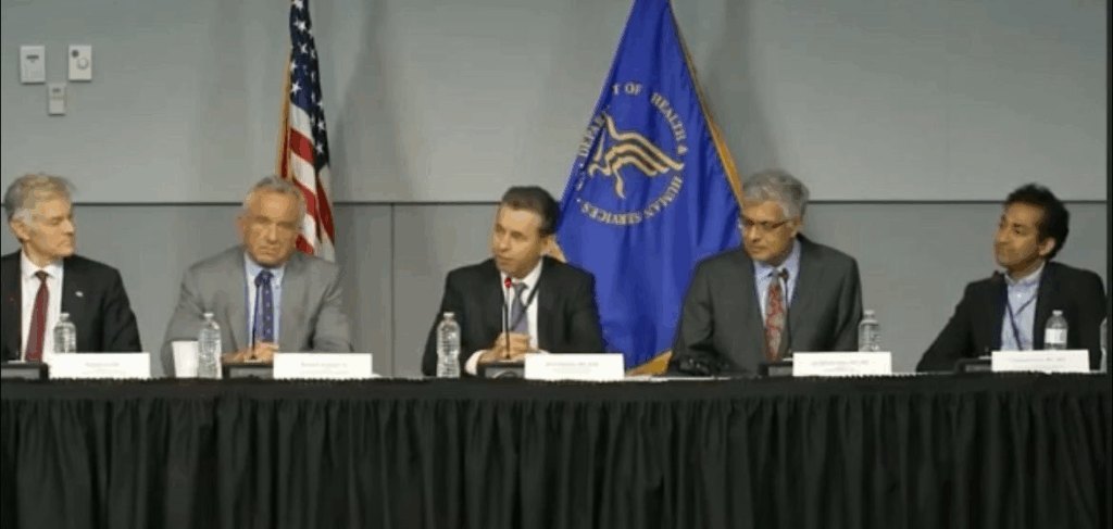 Five men in suits sit at a long table with microphones and nameplates, participating in a panel discussion. An American flag and an official blue government flag are displayed behind them.