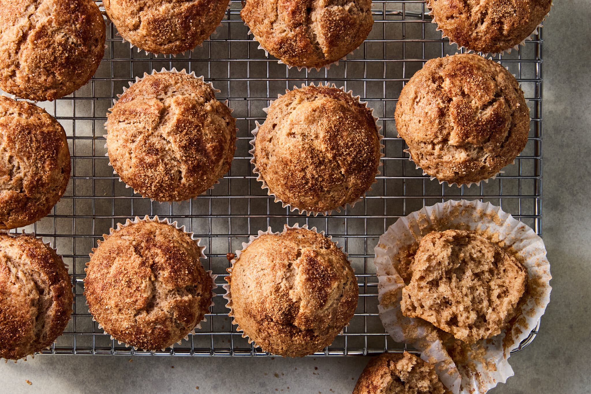 A top-down view of a dozen muffins arranged on a wire rack. One muffin in the bottom right corner has been torn open, revealing its fluffy interior.