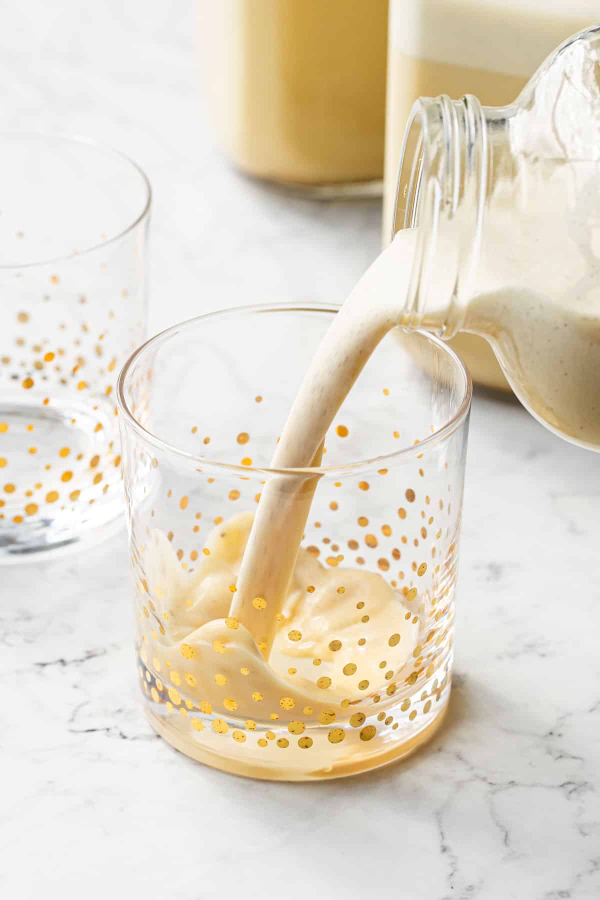 Pouring homemade eggnog into festive gold polka dot glasses on a marble background.