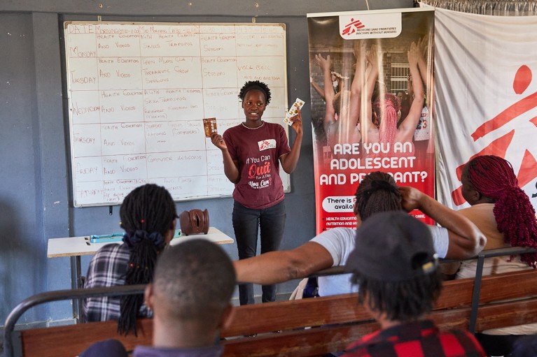 A peer educator stands in front of a whiteboard and Doctors Without Borders banners, showing colleagues two packages.